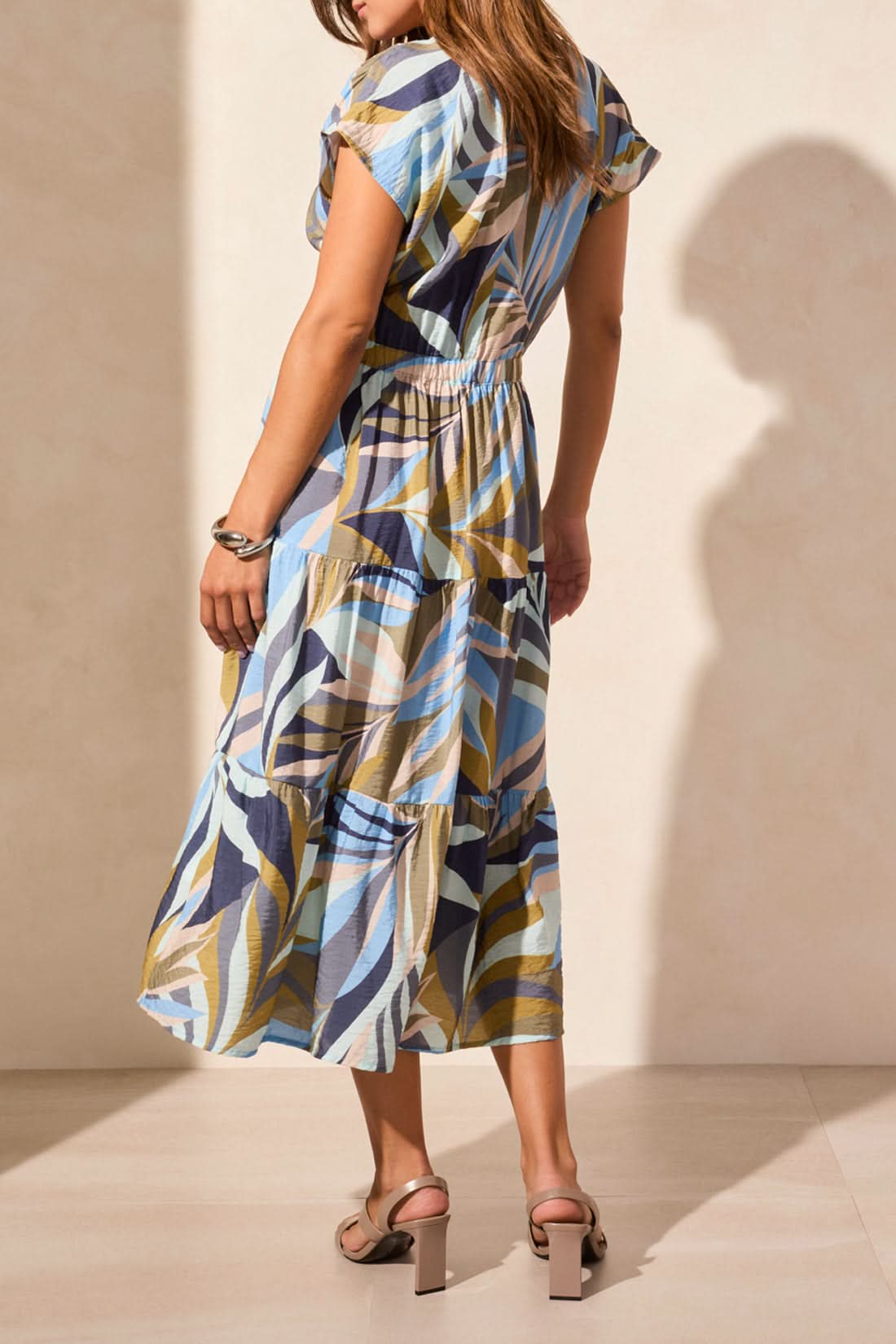 A woman wears the Tribal SHORT SLEEVE V NECK TIERED DRESS with blue, beige, and brown swirls, paired with tan heeled sandals, standing against a light-colored wall.