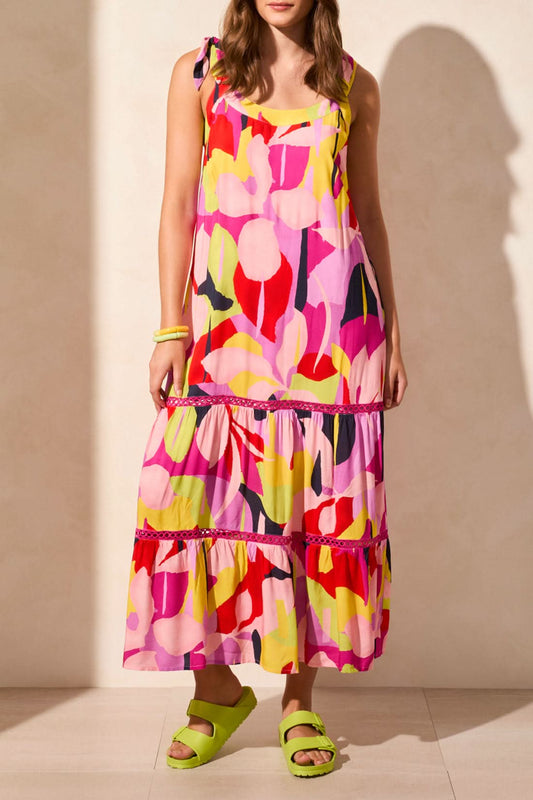 A woman in the Tribal SLEEVLESS DRESS WITH LINING—featuring a colorful abstract print—pairs neon green sandals and matching bracelets as she stands against a light-colored wall, creating a chic summer look.