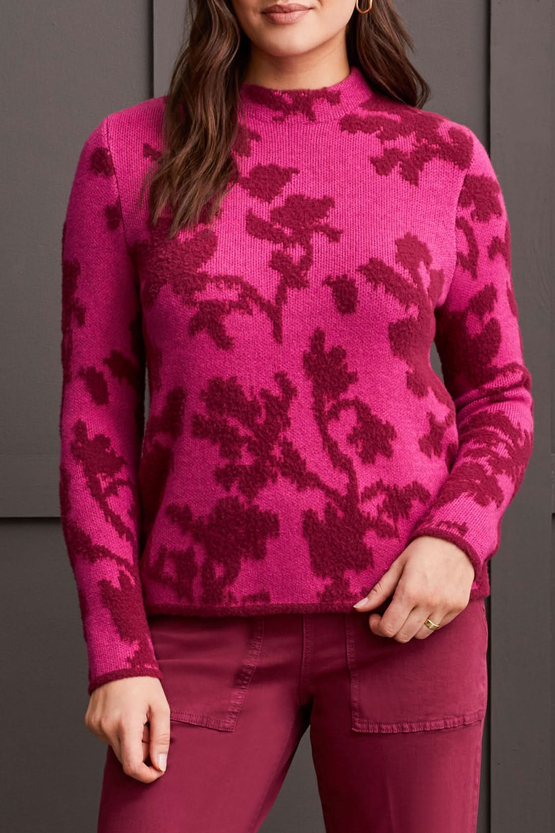 A person dressed in a Tribal Mock Neck Jacquard Sweater featuring dark pink floral patterns and coordinated pink pants stands against a dark background.