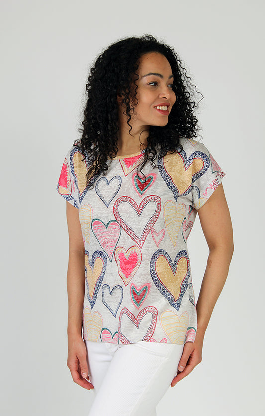 Woman wearing a Fresh FX Hearts Burnout Cap Sleeve Tee with a unique design of heart patterns and white pants, smiling and looking to the side.