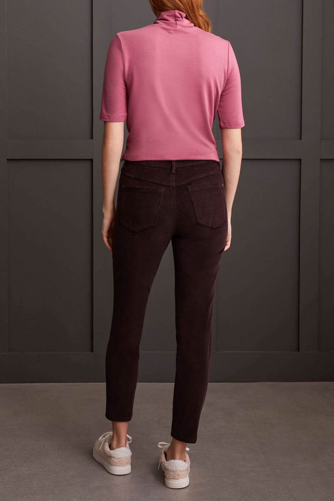 A person clad in a cream-colored quilted coat, a striped top, Tribal Audrey Pull on Slim Ankle Pants in burgundy, and white sneakers stands against a paneled grey wall.