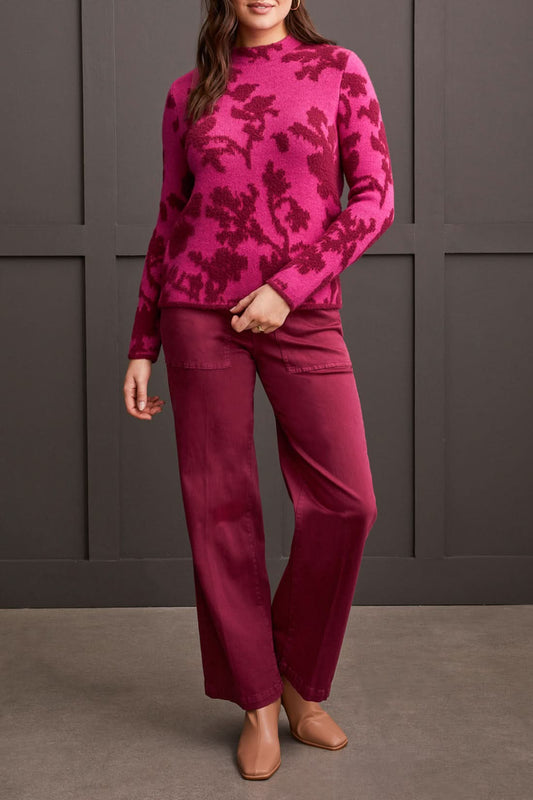 A person dressed in a Tribal Mock Neck Jacquard Sweater featuring dark pink floral patterns and coordinated pink pants stands against a dark background.