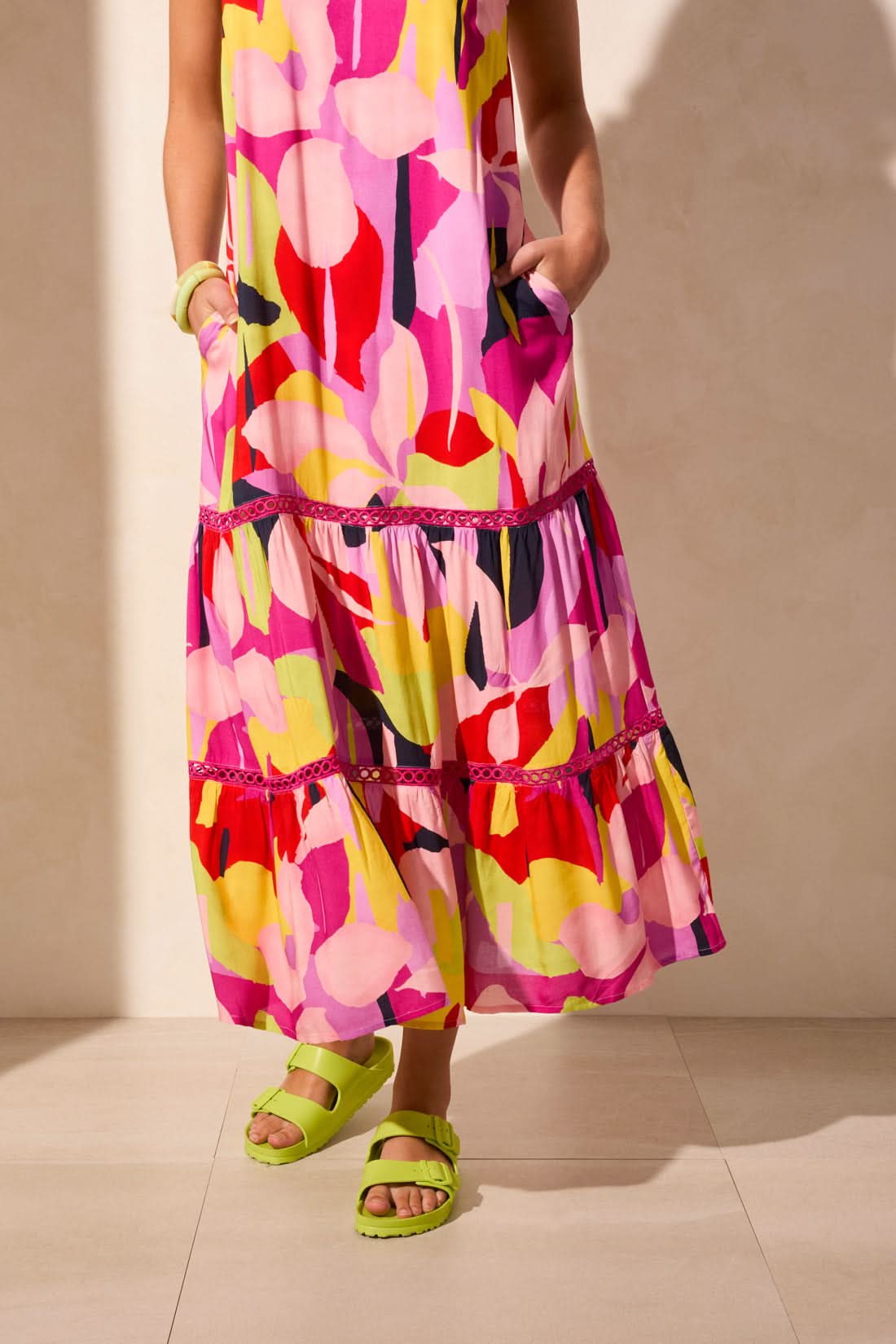 A woman in the Tribal SLEEVLESS DRESS WITH LINING—featuring a colorful abstract print—pairs neon green sandals and matching bracelets as she stands against a light-colored wall, creating a chic summer look.