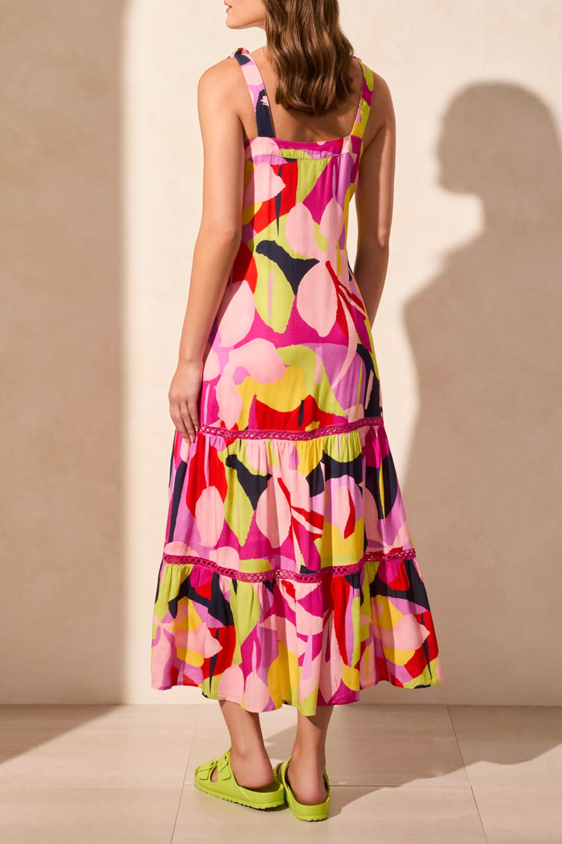 A woman in the Tribal SLEEVLESS DRESS WITH LINING—featuring a colorful abstract print—pairs neon green sandals and matching bracelets as she stands against a light-colored wall, creating a chic summer look.