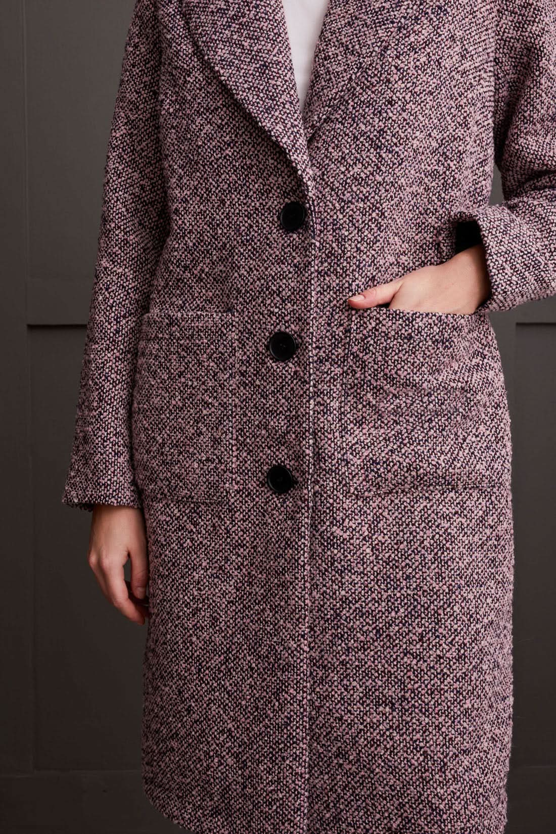 A person wearing a cozy Unlined Button Up Duster Coat by Tribal over a white shirt, pinkish pants, and beige shoes stands with hands in pockets in front of a dark paneled wall.