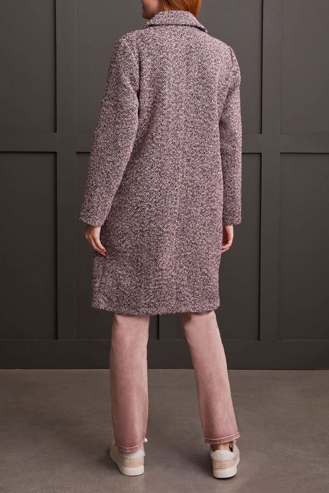 A person wearing a cozy Unlined Button Up Duster Coat by Tribal over a white shirt, pinkish pants, and beige shoes stands with hands in pockets in front of a dark paneled wall.