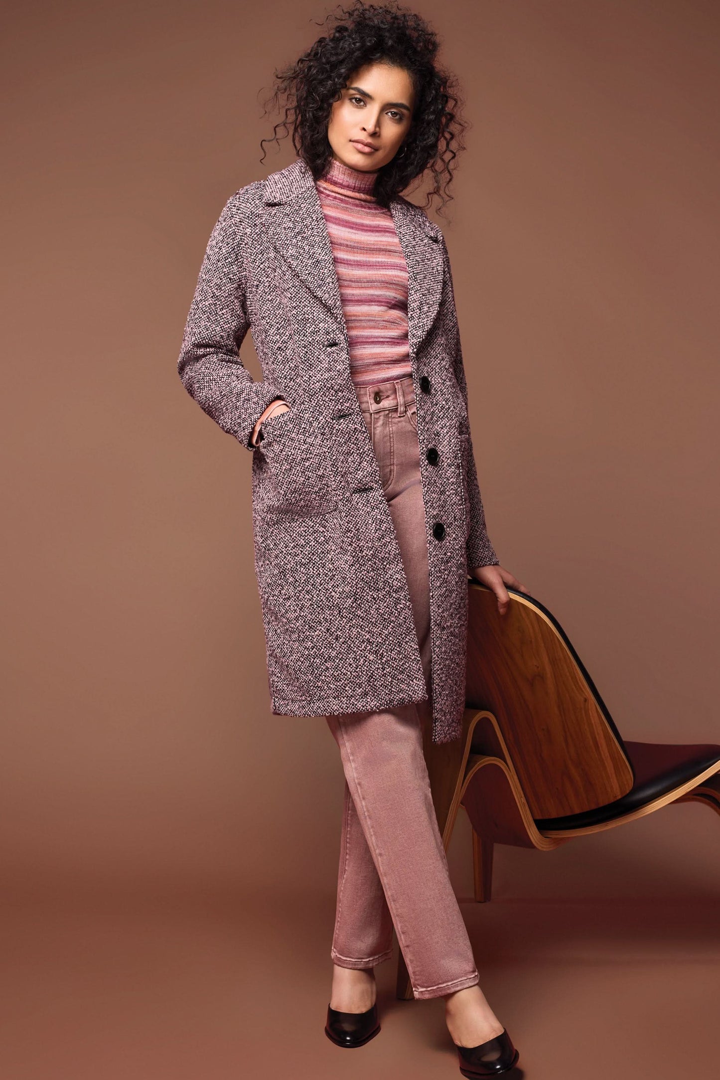A person wearing a cozy Unlined Button Up Duster Coat by Tribal over a white shirt, pinkish pants, and beige shoes stands with hands in pockets in front of a dark paneled wall.
