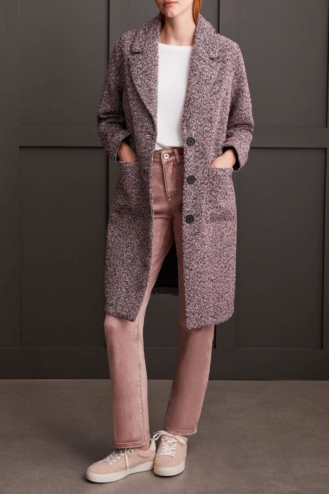 A person wearing a cozy Unlined Button Up Duster Coat by Tribal over a white shirt, pinkish pants, and beige shoes stands with hands in pockets in front of a dark paneled wall.