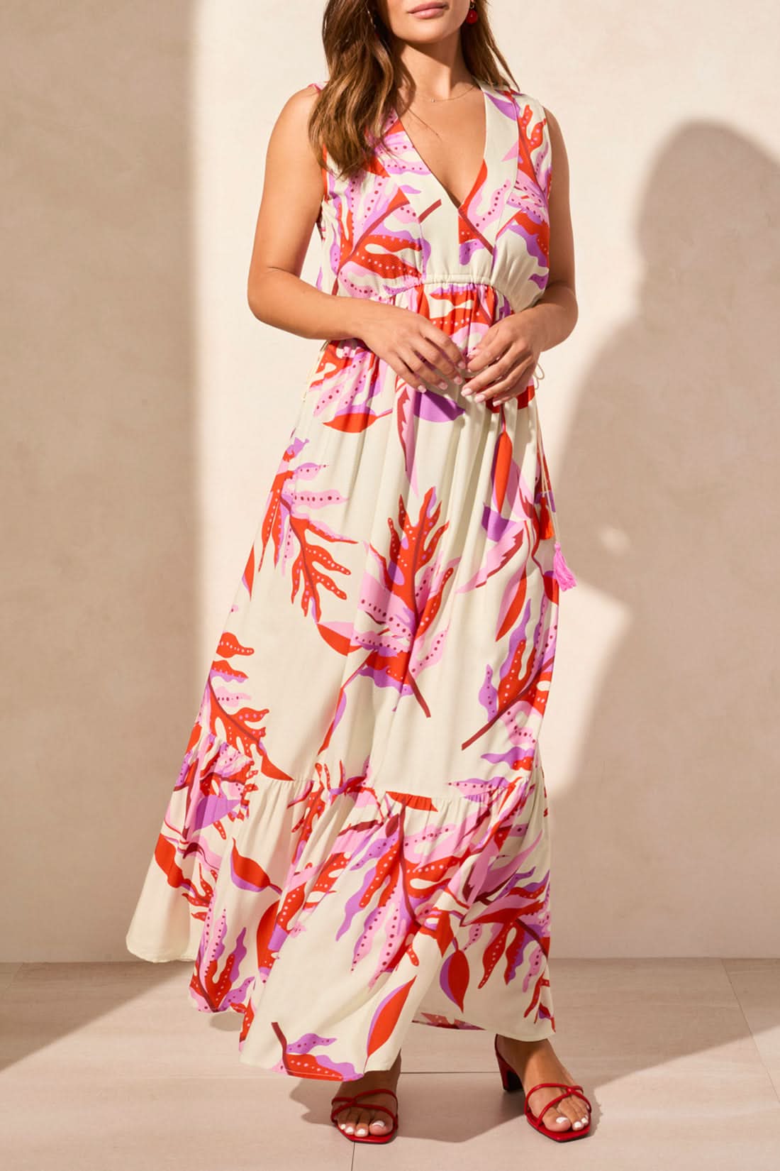 A woman models the Tribal MAXI DRESS WITH WAIST DRAWCORD, a sleeveless v-neck style with a pink and red abstract print, paired with red sandals—an effortless summer wardrobe addition—against a plain light backdrop.