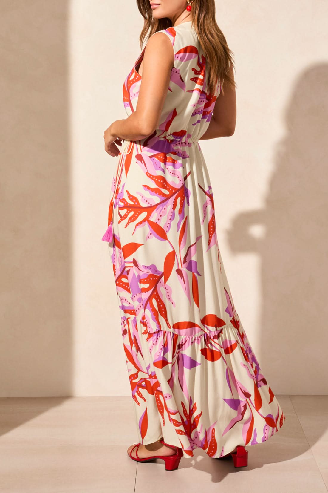 A woman models the Tribal MAXI DRESS WITH WAIST DRAWCORD, a sleeveless v-neck style with a pink and red abstract print, paired with red sandals—an effortless summer wardrobe addition—against a plain light backdrop.