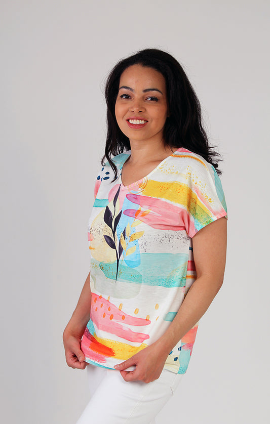 A smiling woman wearing a Fresh FX watercolor cap sleeve top and white pants standing against a plain background.