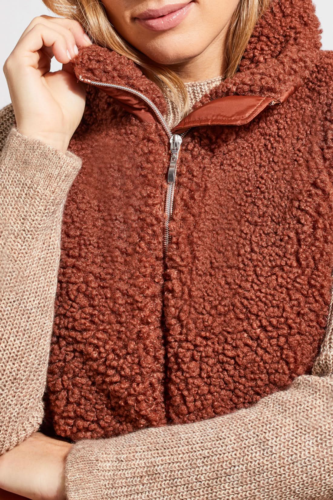 A person stands wearing a beige sweater, blue jeans, brown ankle boots, and a Tribal Sleeveless Sherpa Combo Nylon Vest in brown. The background is plain and white.