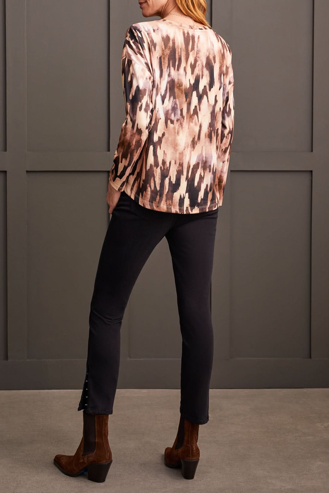 A person wearing the Tribal Crew Neck Top with Curved Hem, featuring a stylish patterned design, paired with a black knee-length skirt and black ankle boots, stands against a dark paneled wall—perfect for any fall wardrobe.
