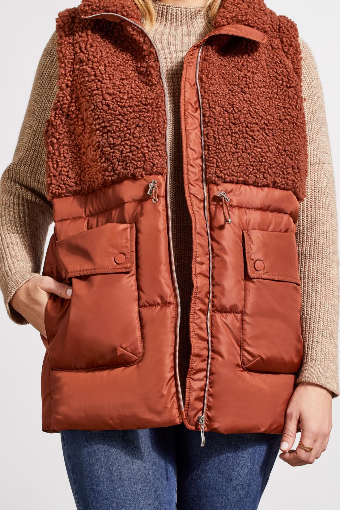 A person stands wearing a beige sweater, blue jeans, brown ankle boots, and a Tribal Sleeveless Sherpa Combo Nylon Vest in brown. The background is plain and white.