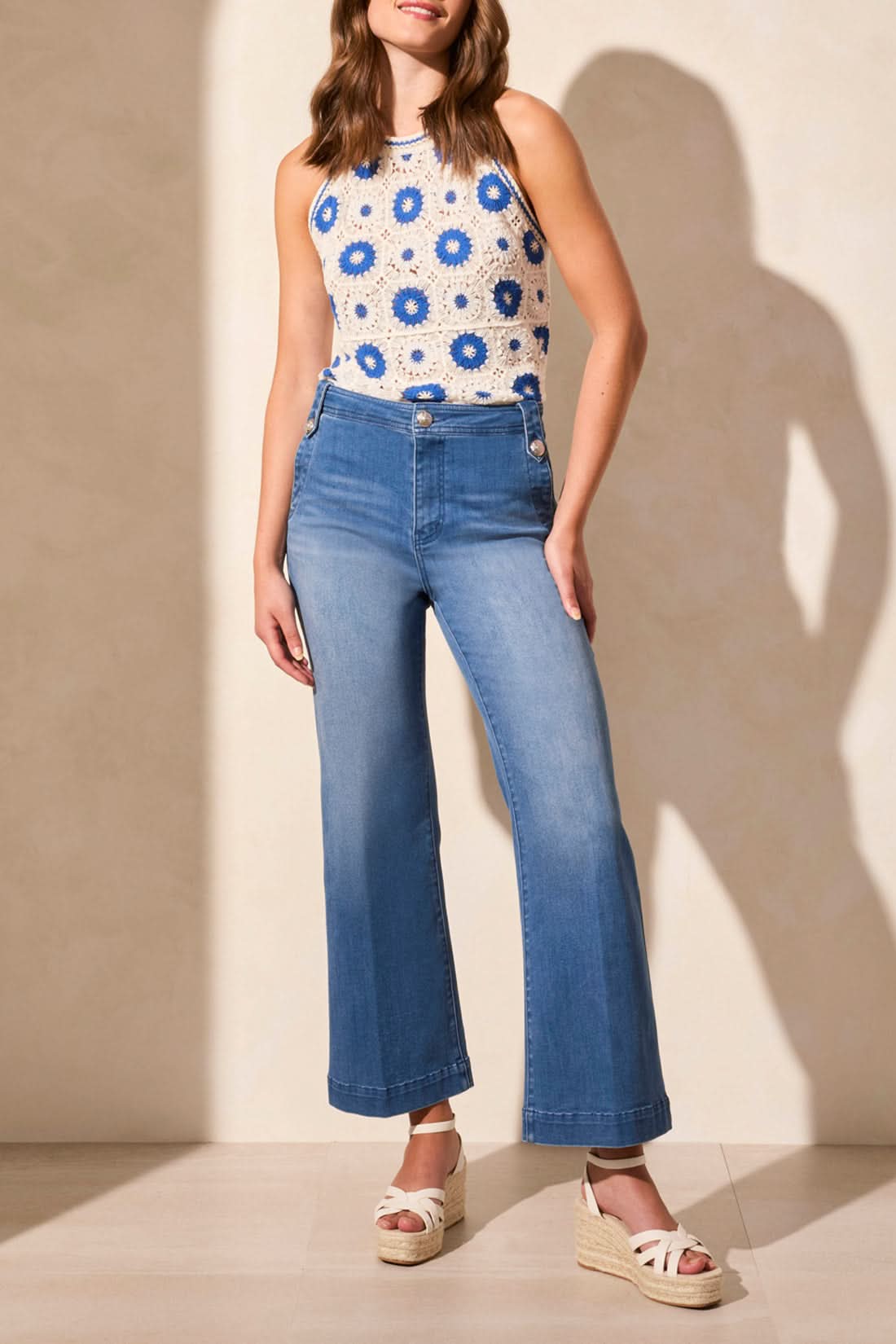 A person wearing a white and blue floral sleeveless top, Tribal’s BROOKE HUGGING PALAZZO MARINE JEANS, and white wedge sandals stands against a light-colored wall.