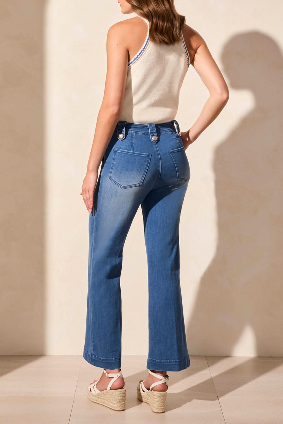 A person wearing a white and blue floral sleeveless top, Tribal’s BROOKE HUGGING PALAZZO MARINE JEANS, and white wedge sandals stands against a light-colored wall.