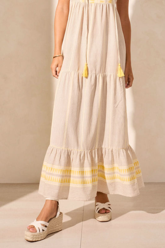 A woman in the Tribal SUMMER VACATION DRESS stands indoors by a wooden table with white vases, casting a shadow—an ideal pick to brighten your summer wardrobe or make vacation days special.