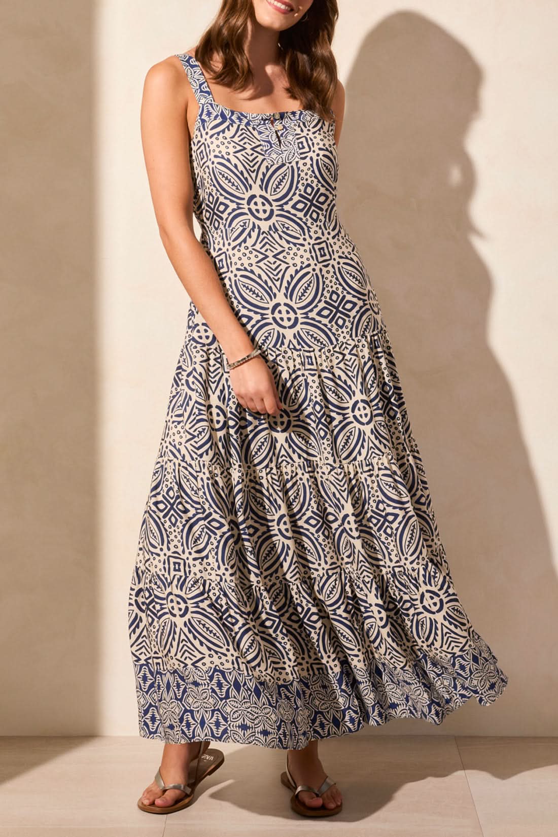 A woman wears the Tribal Sleeveless Tiered Maxi Dress with Lining, featuring a blue and white geometric print. The flowy design offers movement, while its breathable fabric ensures all-day comfort. She stands against a neutral background in sandals.
