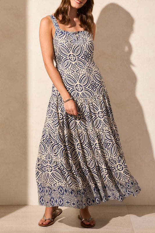 A woman wears the Tribal Sleeveless Tiered Maxi Dress with Lining, featuring a blue and white geometric print. The flowy design offers movement, while its breathable fabric ensures all-day comfort. She stands against a neutral background in sandals.