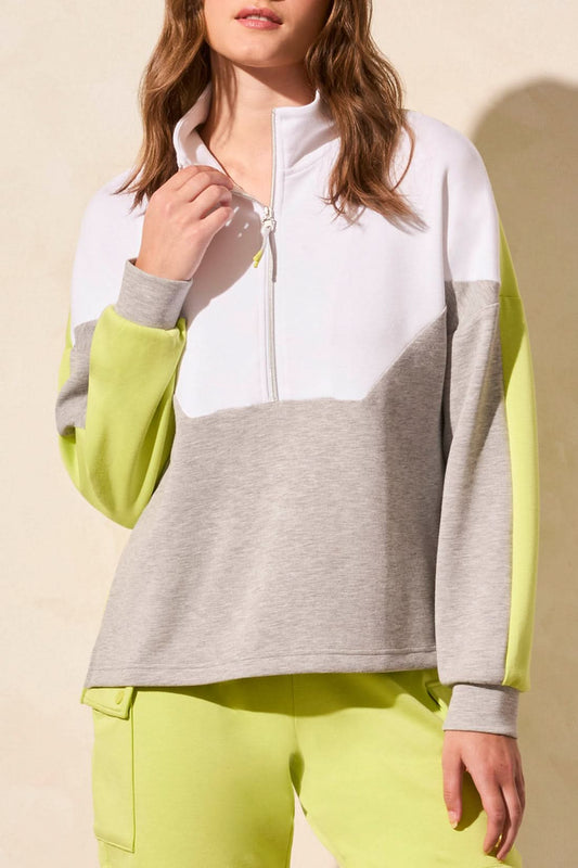 A woman wears the Tribal LONG SLEEVE MOCK NECK 1/4 ZIP top, featuring a white zip collar, gray body, and neon yellow sleeves, styled with neon yellow pants. Her face is partially out of frame.