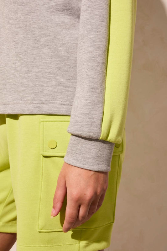 A woman wears the Tribal LONG SLEEVE MOCK NECK 1/4 ZIP top, featuring a white zip collar, gray body, and neon yellow sleeves, styled with neon yellow pants. Her face is partially out of frame.