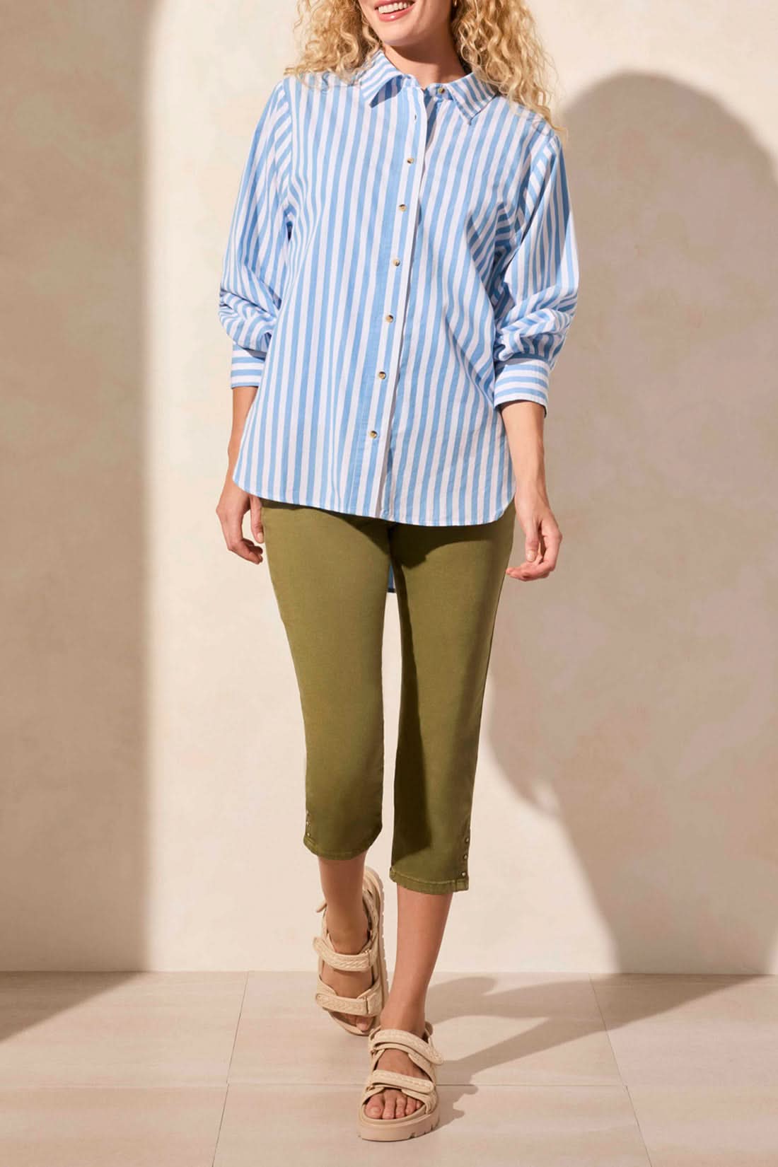 A woman models the Tribal LONG SLEEVE BUTTON FRONT SHIRT in blue and white vertical stripes with rolled sleeves, paired with olive green pants. Her face is partially out of frame for a relaxed yet stylish look.