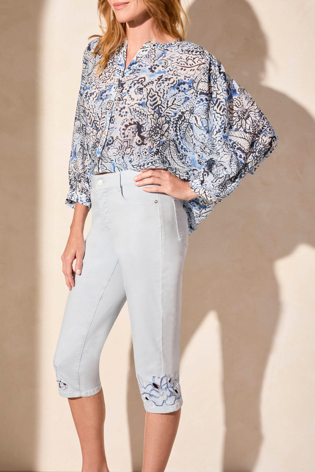 Woman wearing a blue patterned blouse and Tribal SOPHIA CURVY CAPRI JEANS—stylish capris with embroidered cuffs—paired with beige heeled sandals, standing against a plain light background.