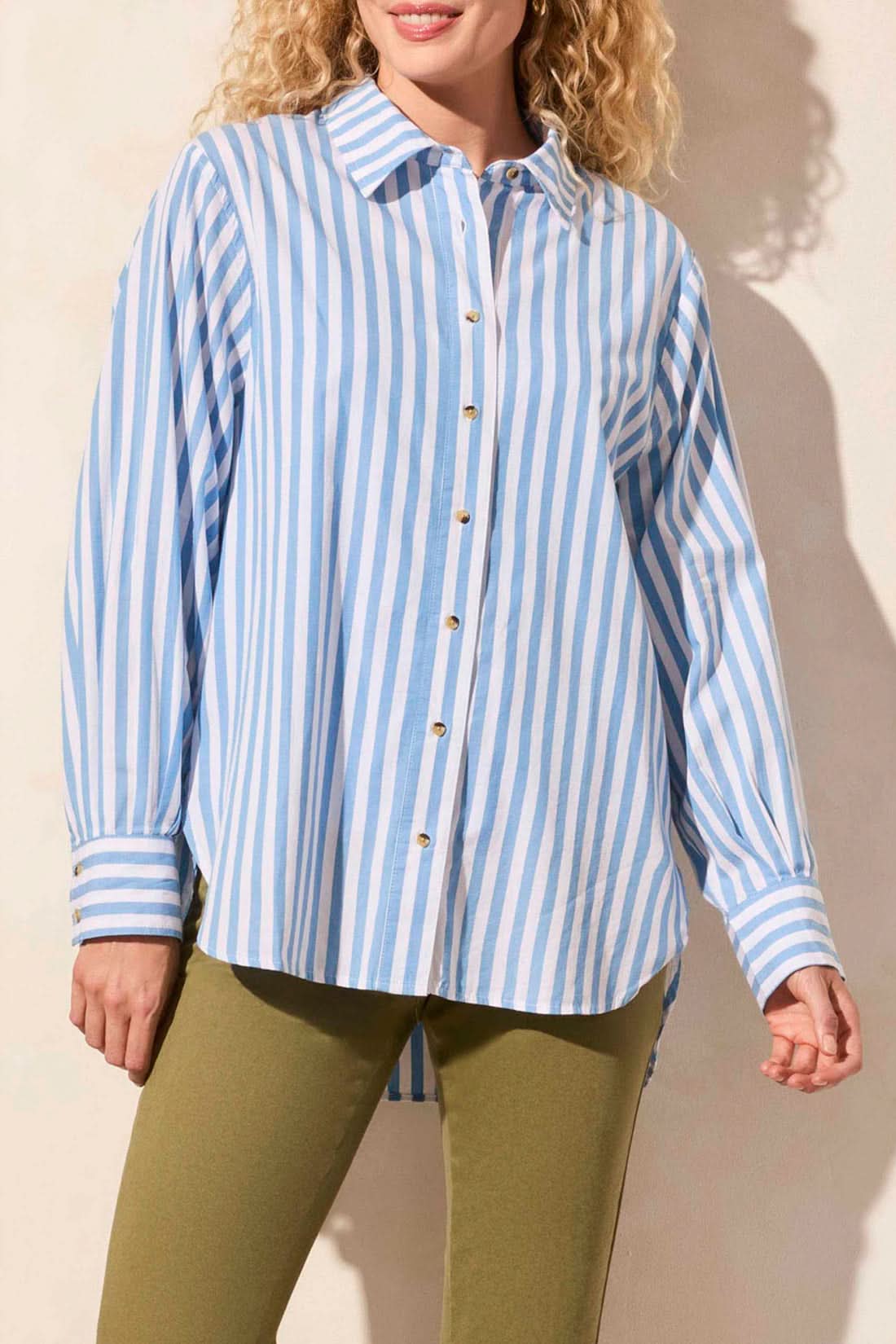 A woman models the Tribal LONG SLEEVE BUTTON FRONT SHIRT in blue and white vertical stripes with rolled sleeves, paired with olive green pants. Her face is partially out of frame for a relaxed yet stylish look.