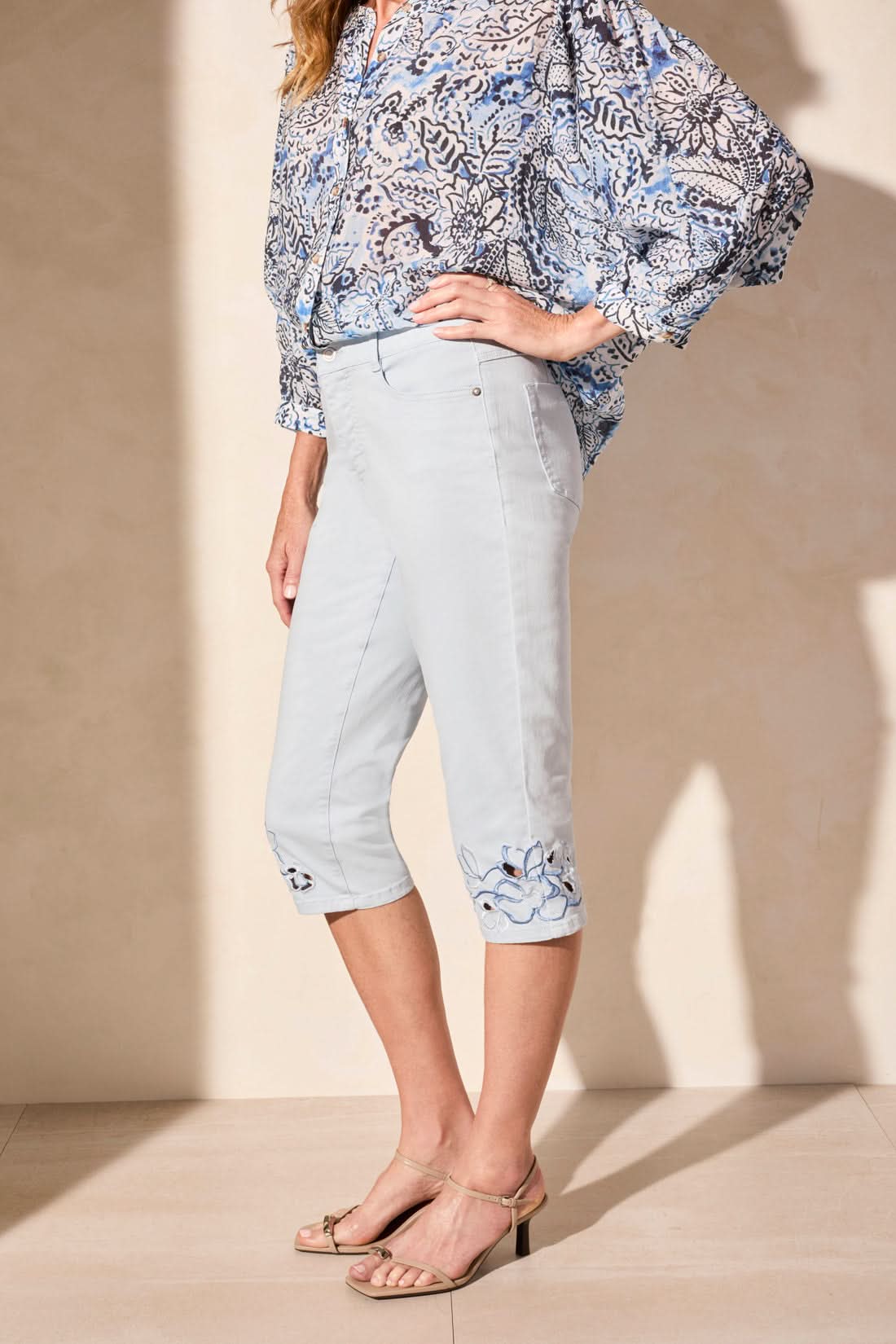 Woman wearing a blue patterned blouse and Tribal SOPHIA CURVY CAPRI JEANS—stylish capris with embroidered cuffs—paired with beige heeled sandals, standing against a plain light background.