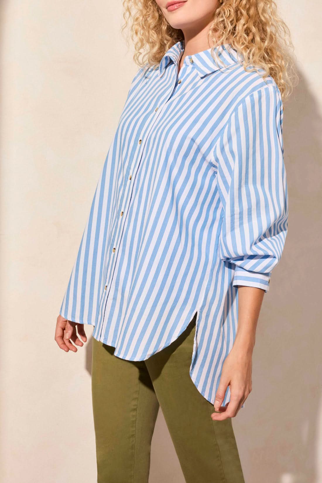 A woman models the Tribal LONG SLEEVE BUTTON FRONT SHIRT in blue and white vertical stripes with rolled sleeves, paired with olive green pants. Her face is partially out of frame for a relaxed yet stylish look.