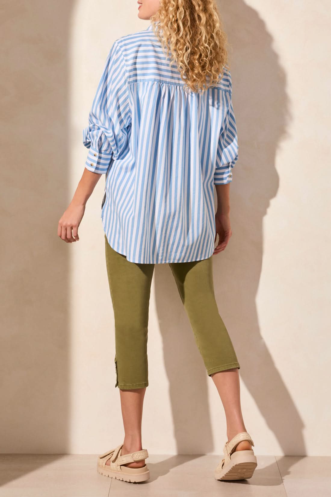A woman models the Tribal LONG SLEEVE BUTTON FRONT SHIRT in blue and white vertical stripes with rolled sleeves, paired with olive green pants. Her face is partially out of frame for a relaxed yet stylish look.