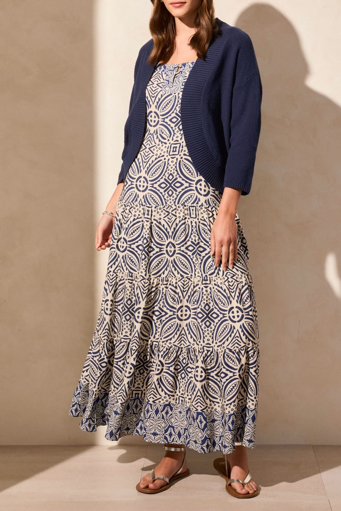 A woman stands facing away, wearing the Tribal COCOON SWEATER CARDIGAN for cozy comfort, paired with a long blue and white geometric patterned skirt and brown sandals.