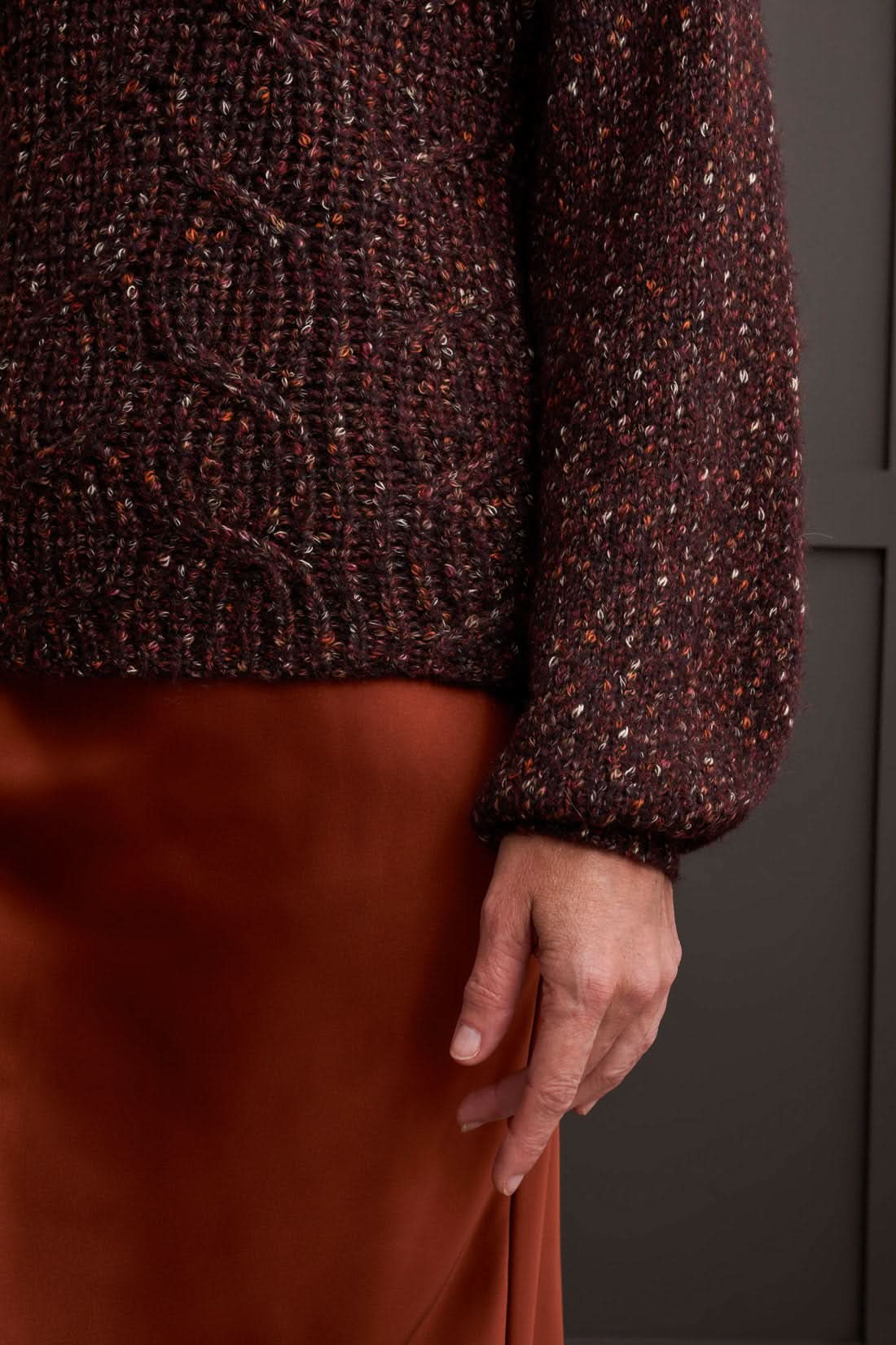 Person wearing a red patterned coat, cozy Tribal Funnel Neck Oversize Sweater in red, blue jeans, and black ankle boots stands against a dark paneled wall.