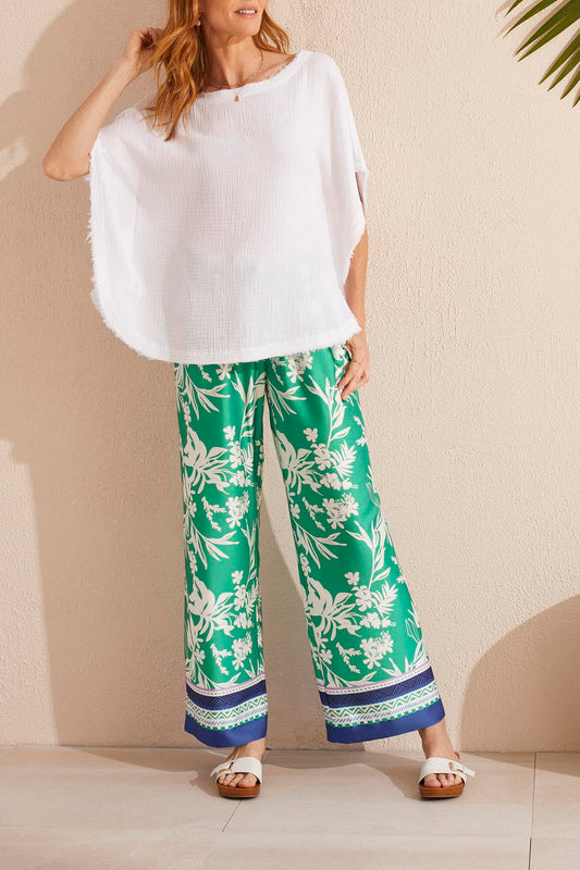 Woman standing against a wall wearing an oversized Tribal white kaftan blouse and green floral pants, one hand adjusting her hair, sunlight casting shadows.
