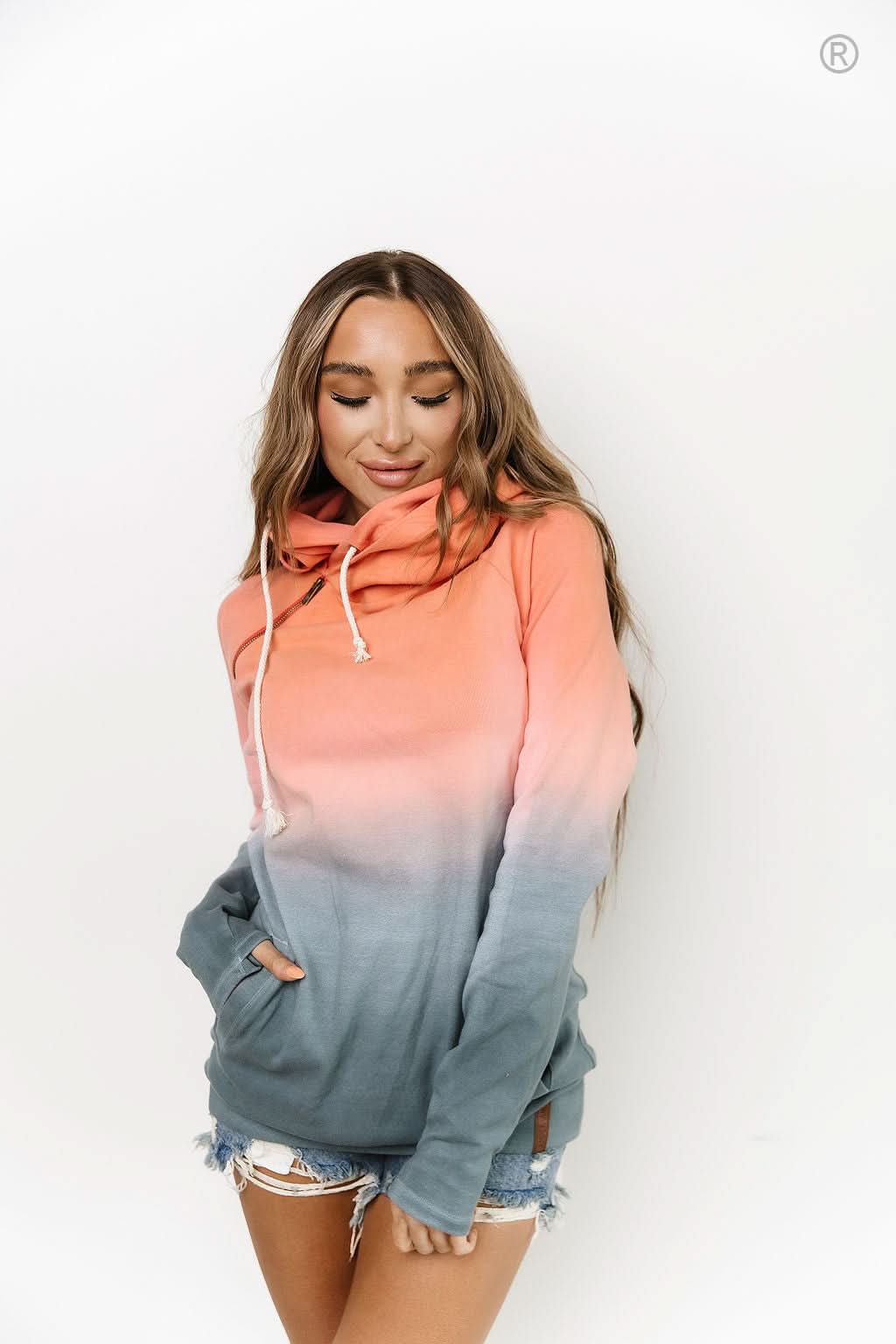 Dressed in Ampersand Avenue's Chasing Sunset Doublehooded Sweatshirt and denim shorts, a person exudes comfort and style while standing in a kitchen adorned with a marble backsplash and wooden cabinets.