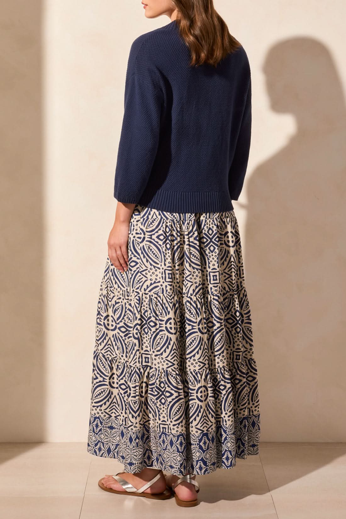 A woman stands facing away, wearing the Tribal COCOON SWEATER CARDIGAN for cozy comfort, paired with a long blue and white geometric patterned skirt and brown sandals.