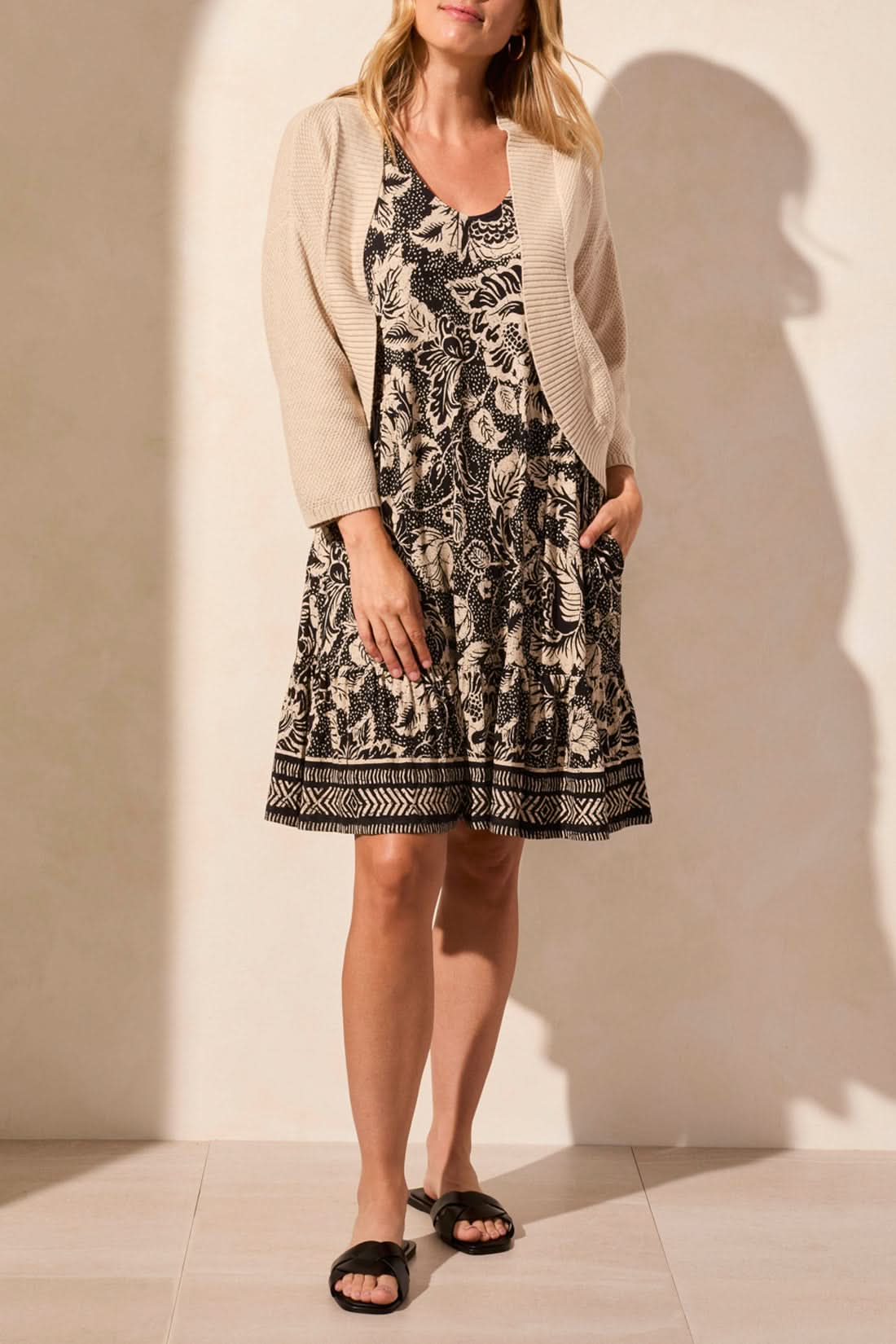 A woman stands facing away, wearing the Tribal COCOON SWEATER CARDIGAN for cozy comfort, paired with a long blue and white geometric patterned skirt and brown sandals.