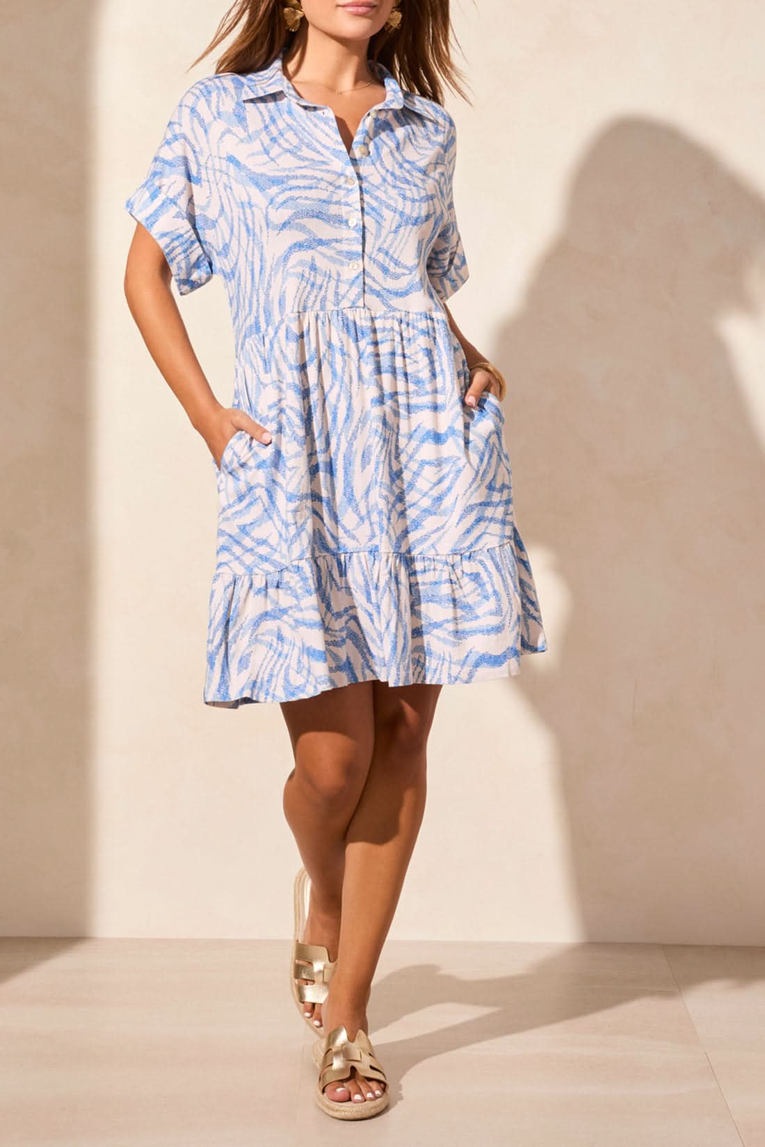 A woman wears the Tribal BUTTON UP DRESS WITH LINING in light blue and white, featuring pockets and a playful pattern, styled with metallic sandals against a beige wall—adding quirky charm to her summer look.