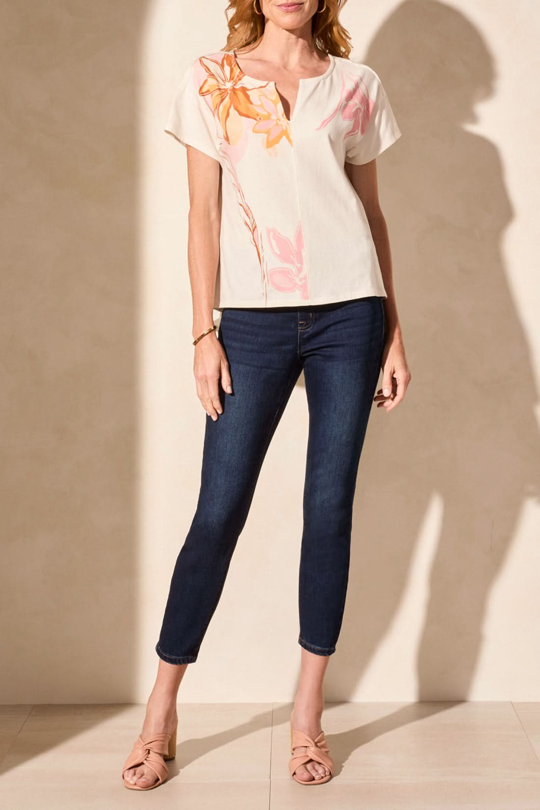A woman stands sideways against a plain background wearing the Tribal SHORT SLEEVE DROP SHOULDER TOP in cream with a pink design, paired with dark skinny jeans and nude heeled sandals for a trendy, relaxed look.