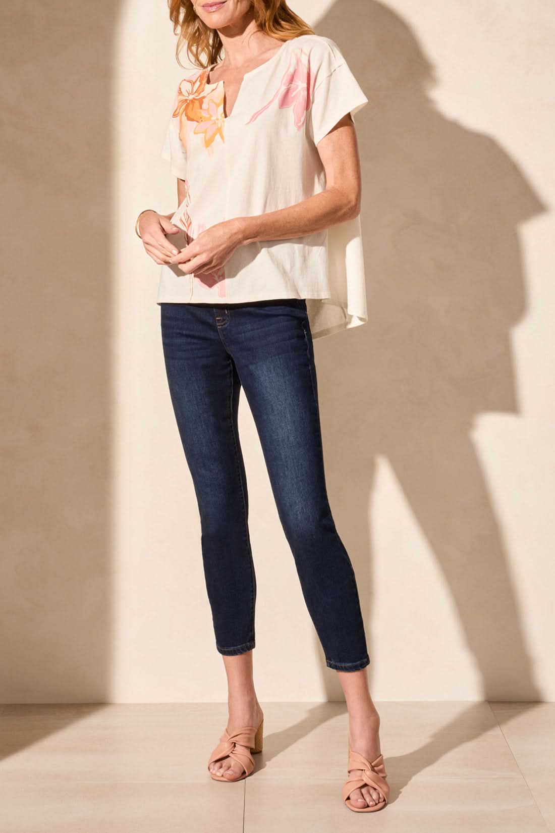 A woman stands sideways against a plain background wearing the Tribal SHORT SLEEVE DROP SHOULDER TOP in cream with a pink design, paired with dark skinny jeans and nude heeled sandals for a trendy, relaxed look.