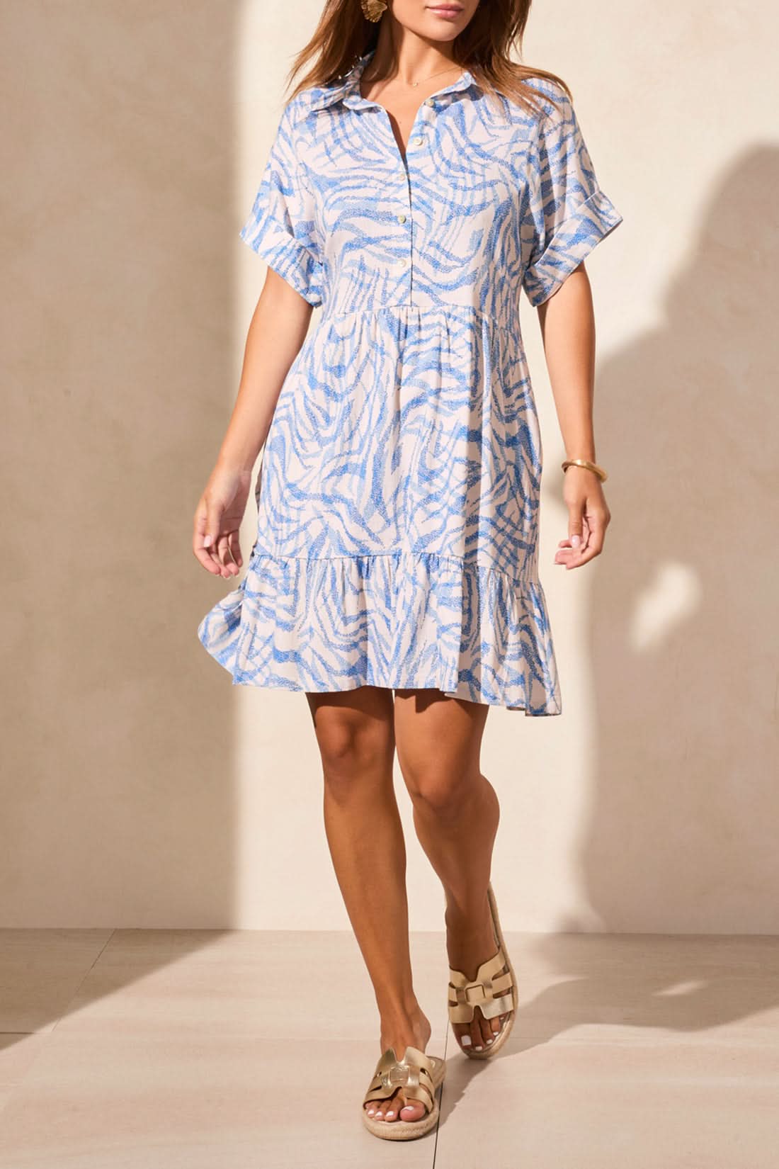 A woman wears the Tribal BUTTON UP DRESS WITH LINING in light blue and white, featuring pockets and a playful pattern, styled with metallic sandals against a beige wall—adding quirky charm to her summer look.