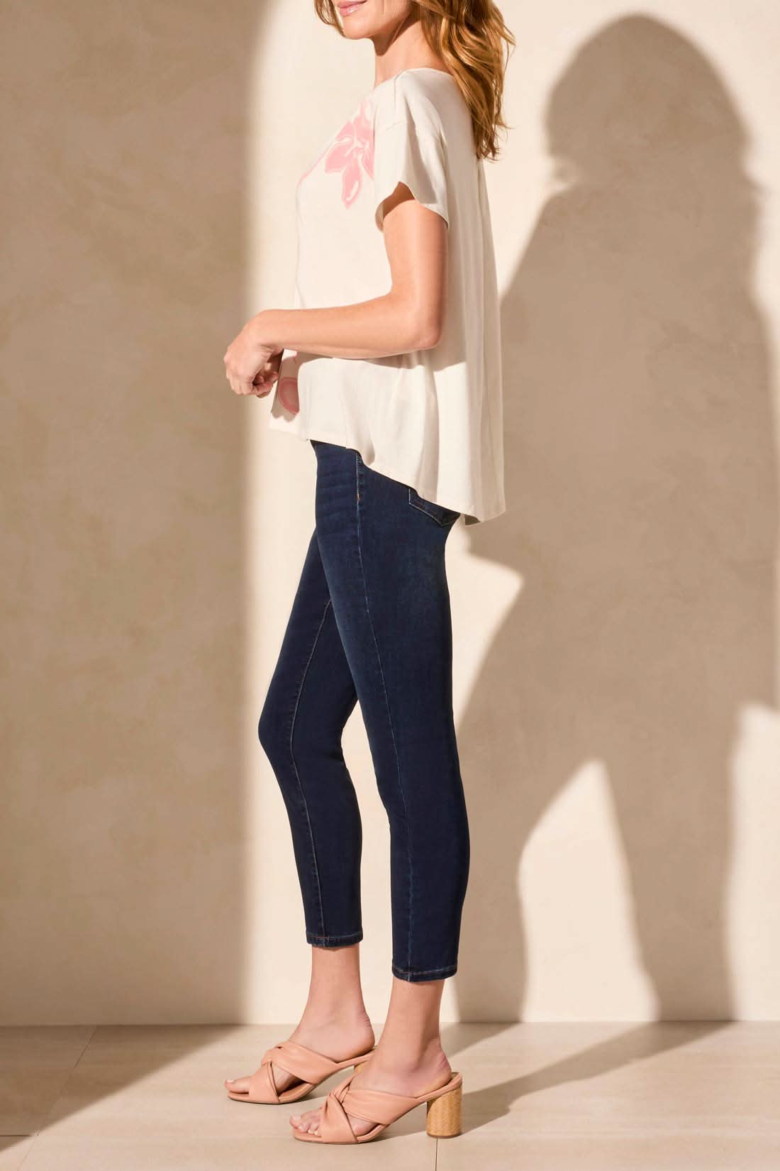 A woman stands sideways against a plain background wearing the Tribal SHORT SLEEVE DROP SHOULDER TOP in cream with a pink design, paired with dark skinny jeans and nude heeled sandals for a trendy, relaxed look.