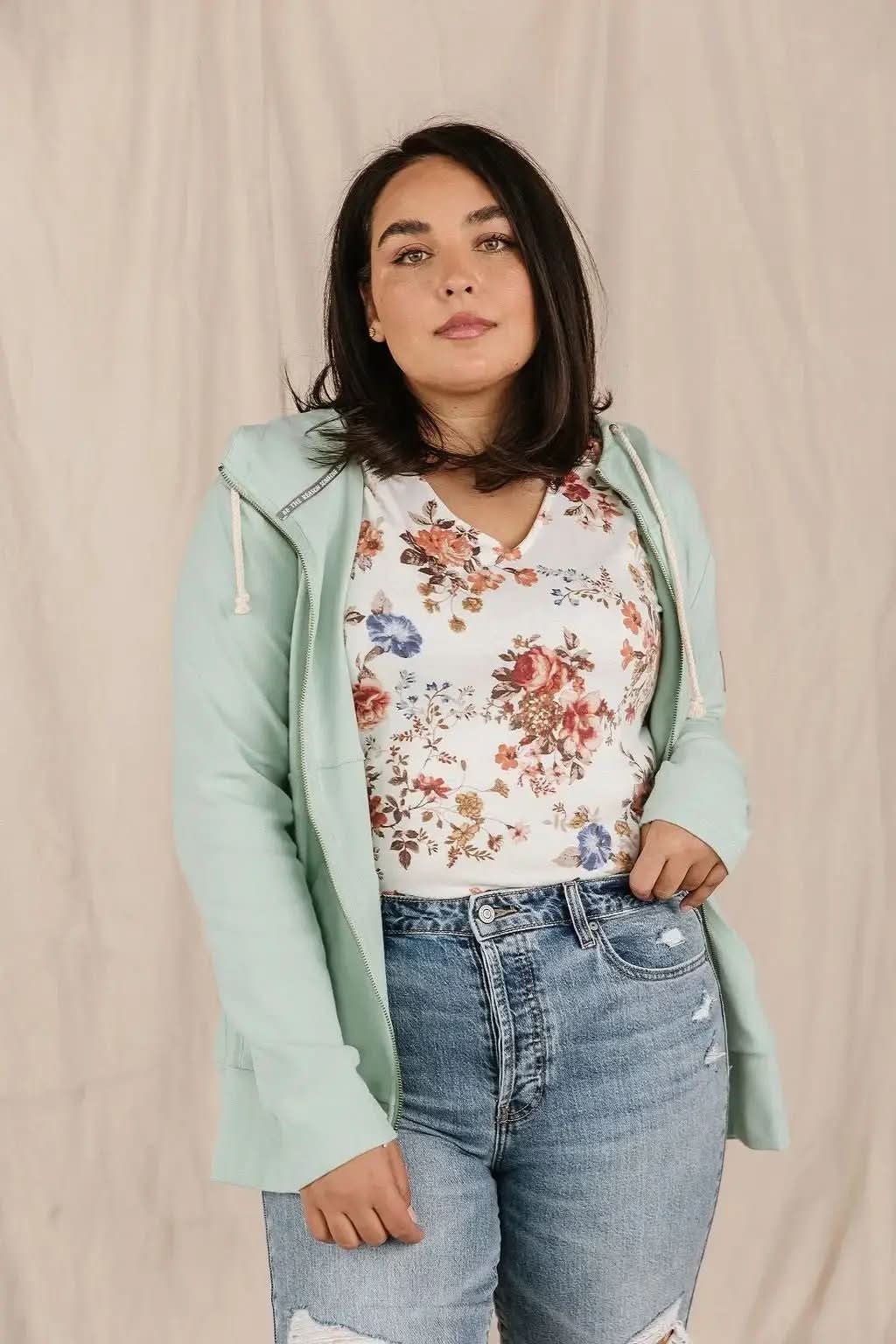 A woman with long hair wearing a UNISHE Seafoam Fullzip Sweatshirt and black shorts stands in front of a mirror, with plants and candles arranged on a table nearby.