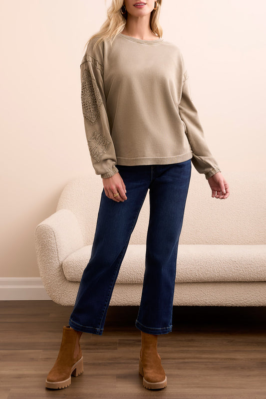 A woman stands indoors wearing the Tribal CREW NECK APPLIQUE DTL sweater, blue cropped jeans, and brown suede ankle boots, with a cream sofa in the background.