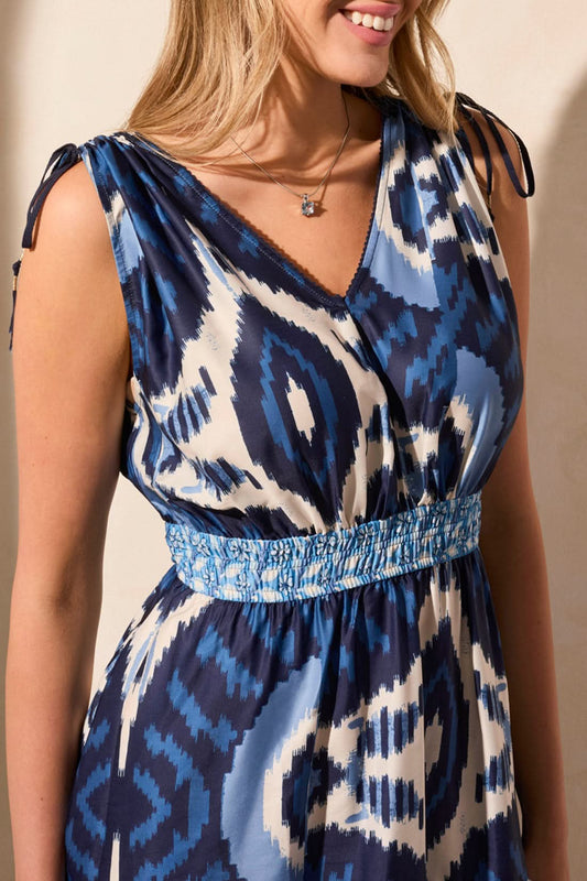 A woman wears the Tribal MAXI DRESS WITH SHOULDER TIES in blue and white, facing away while carrying a beige handbag and sporting white wedge sandals.