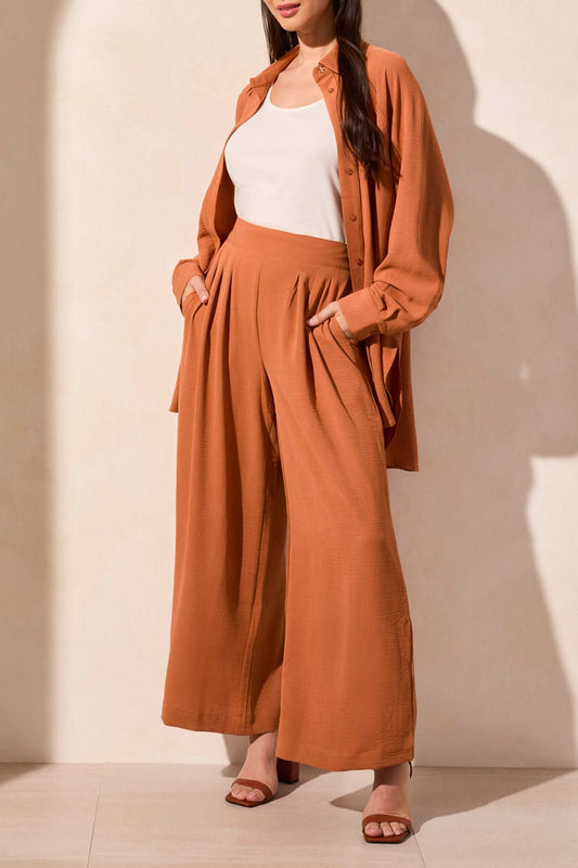 A woman stands against a neutral background wearing a rust-colored button-up shirt, a white top, brown heeled sandals, and Tribal’s FLOWY PANT WITH BLACK ELASTIC WAISTBAND.