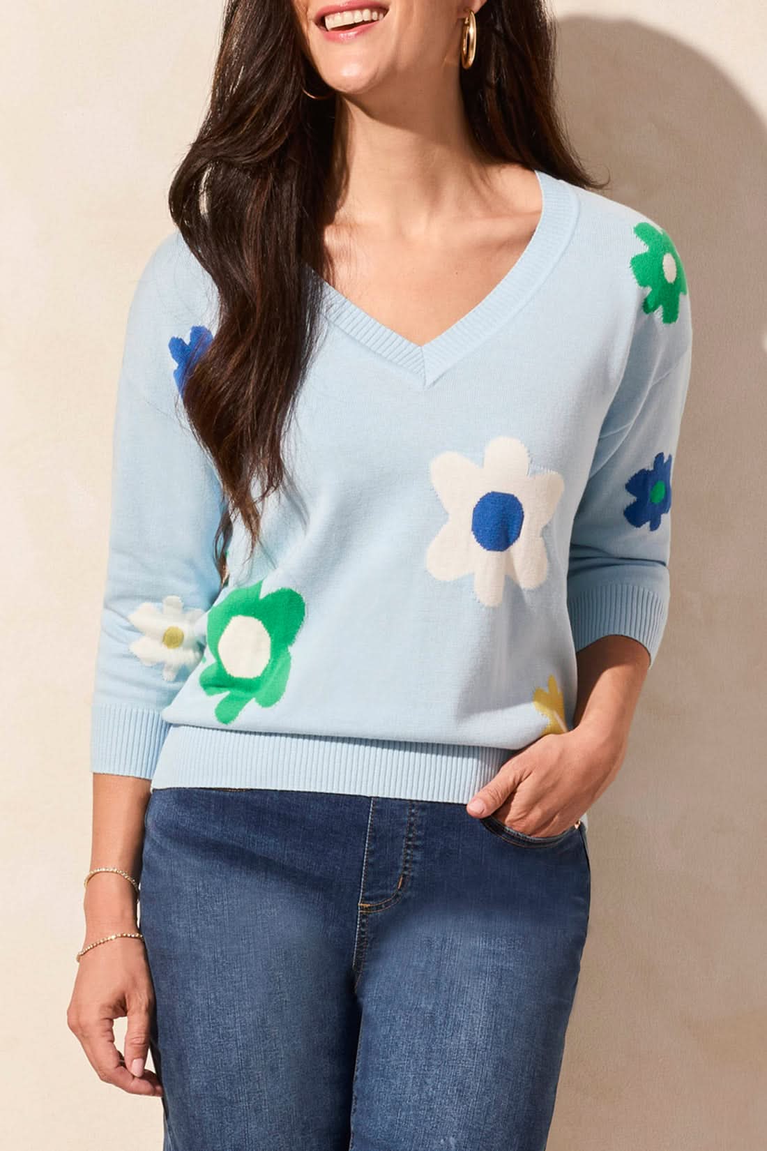 A woman models the Tribal 3/4 Sleeve V-Neck Sweater in light blue with colorful floral patterns, paired with blue jeans and set against a neutral background.
