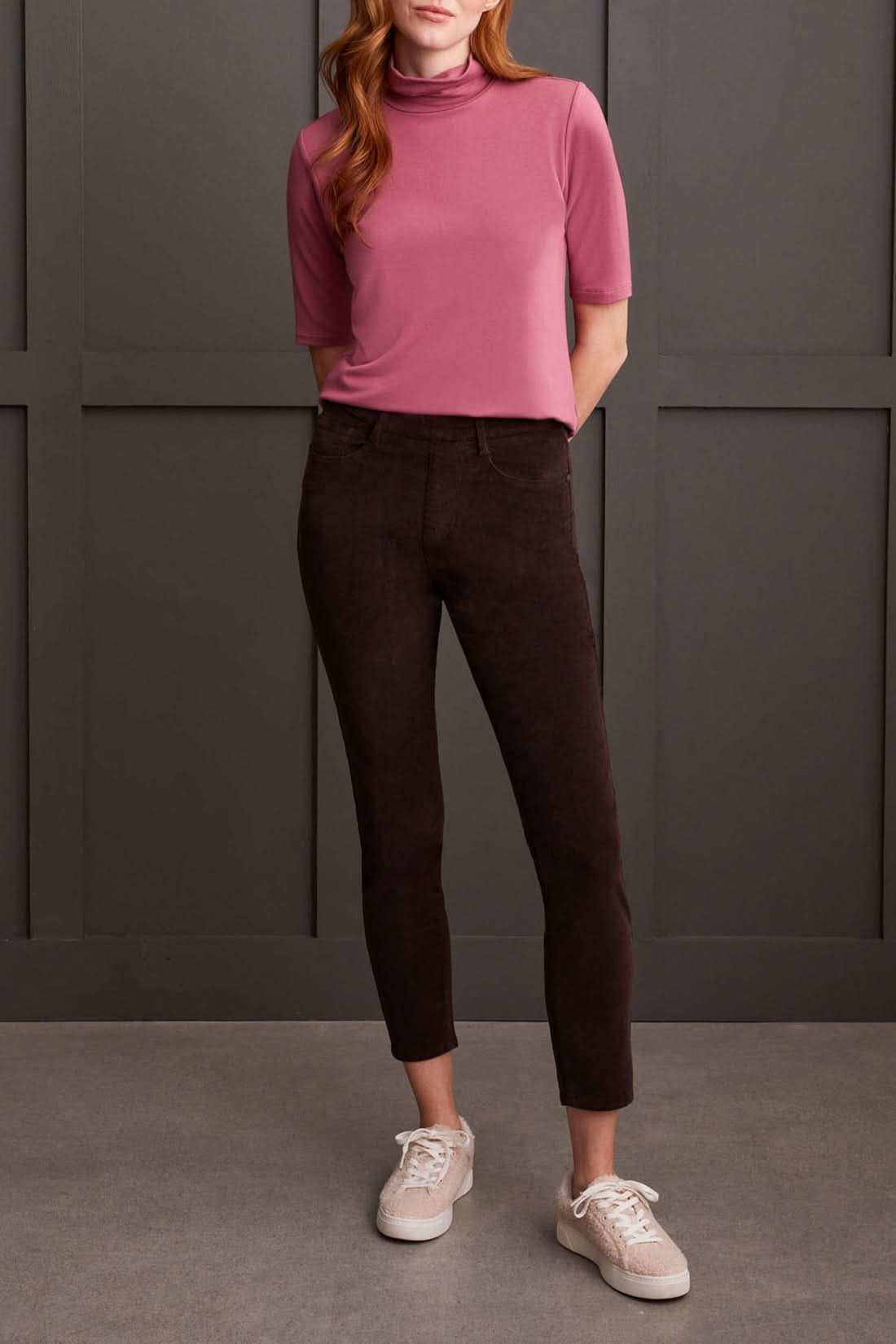 A person clad in a cream-colored quilted coat, a striped top, Tribal Audrey Pull on Slim Ankle Pants in burgundy, and white sneakers stands against a paneled grey wall.