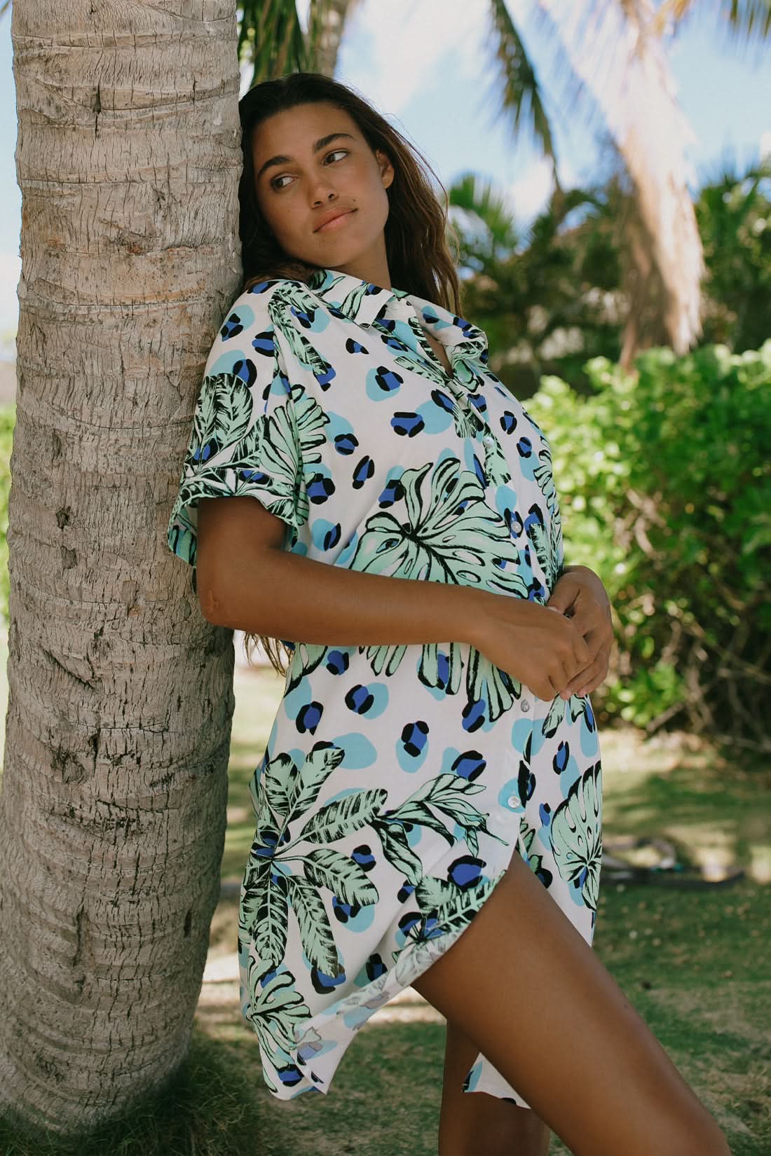 A woman stands outdoors by palm trees, wearing the Tribal SHORT SLEEVE SHIRT DRESS WITH POCKETS in a green and blue tropical print—a must-have for your wardrobe.