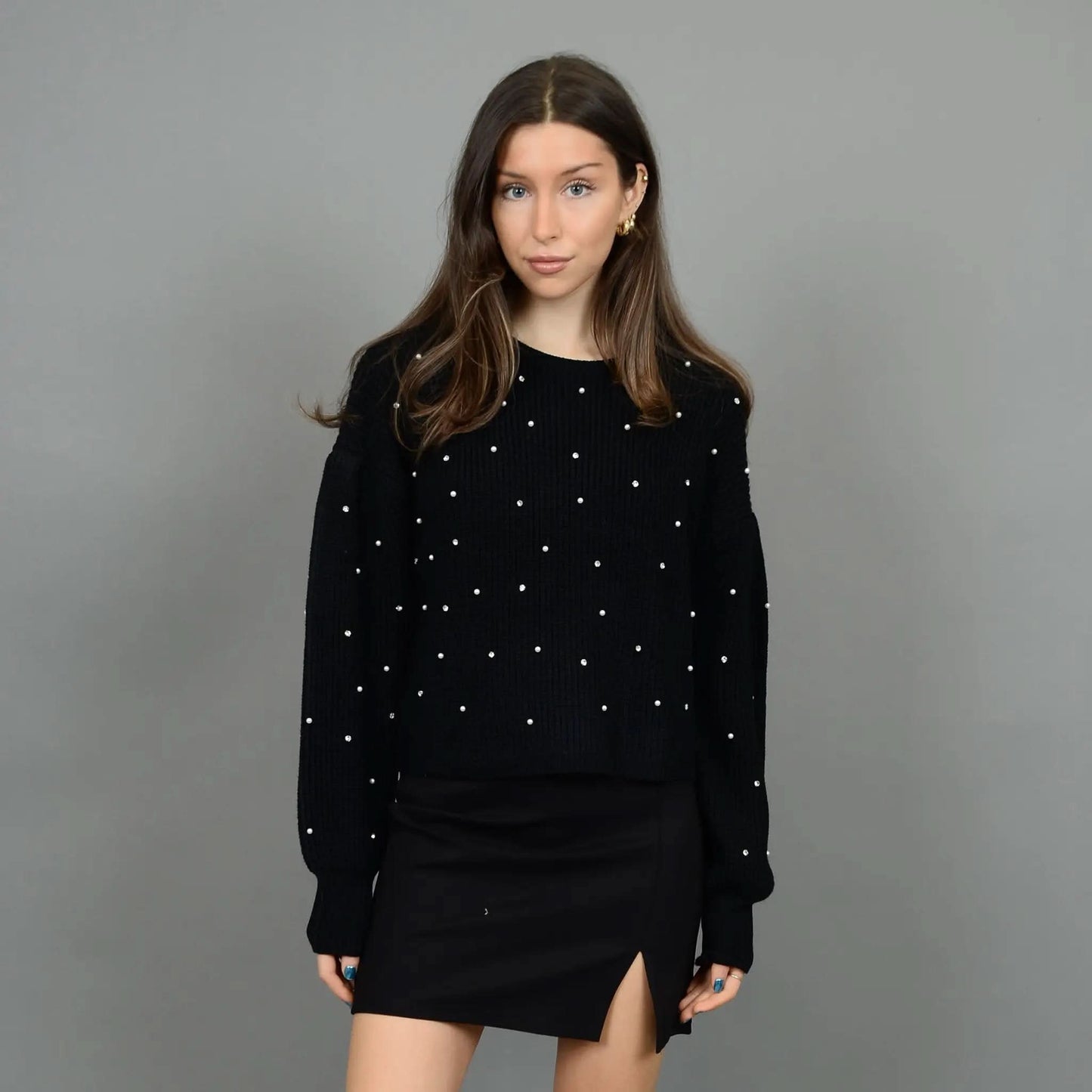 A person stands against a plain background wearing the stylish and cozy RD International Niri Long Sleeve Pull Over Sweater adorned with white dots, paired with a black skirt featuring a front slit. Their hands are in their pockets as they look slightly to their right.